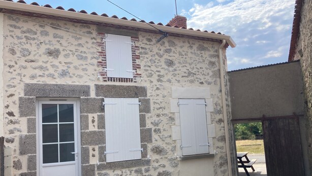A stone building with a white door and shutters, a small patio area with a table and benches, and a clear sky. - Jolie Petite Maison au Cœur du Boccage Vendéen (Le Tablier)