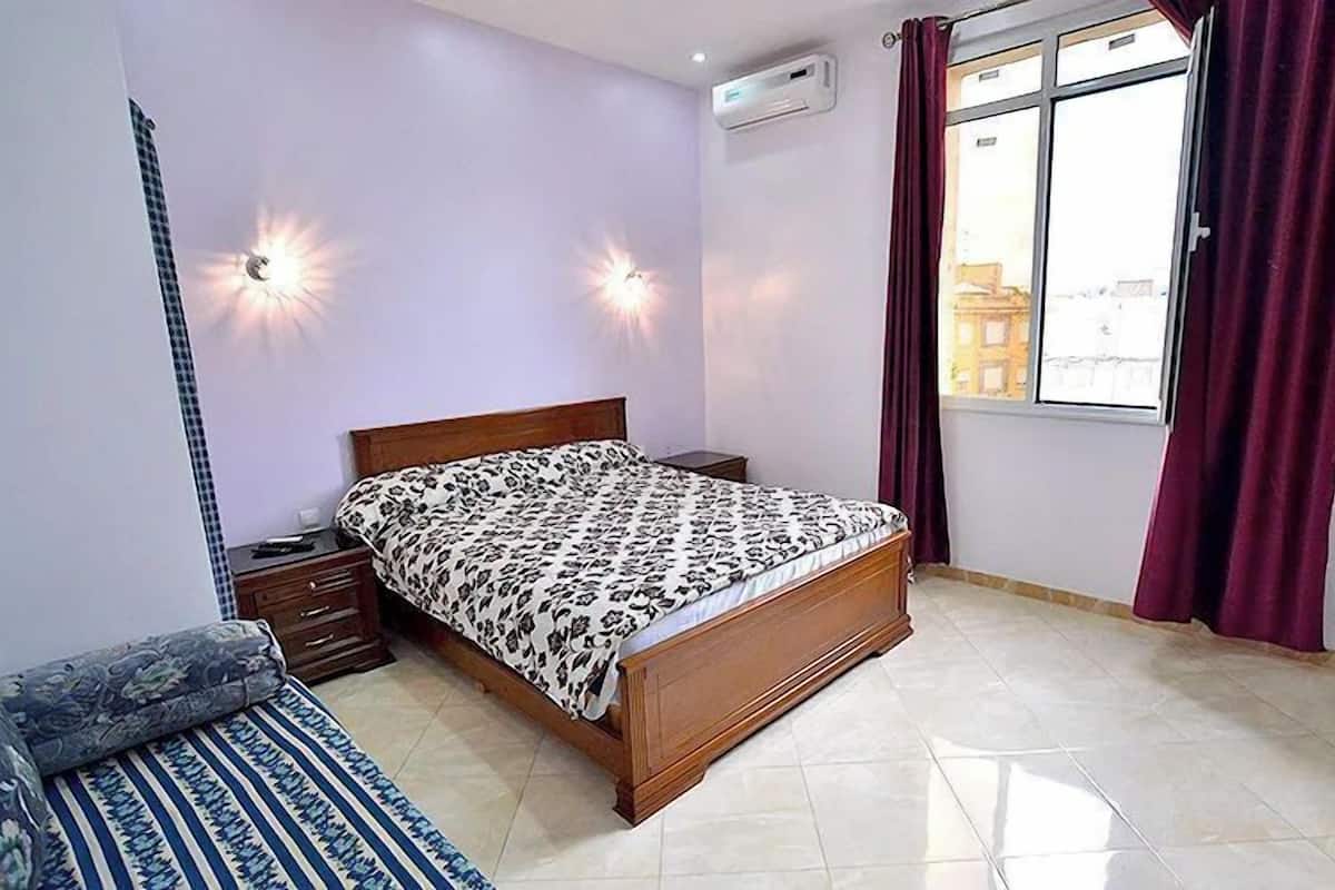 Une chambre à coucher avec un lit en bois, une table de chevet, une lampe, une fenêtre avec des rideaux et un appareil de climatisation.