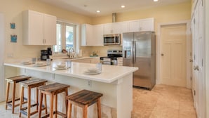 A modern kitchen with white cabinets, stainless steel appliances, and a central island with bar stools. - Castaway Cottage (St. Augustine)
