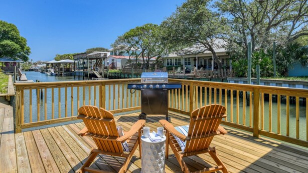 A wooden deck with two wooden chairs and a grill, overlooking a body of water with boats and houses in the background. - Castaway Cottage (St. Augustine)