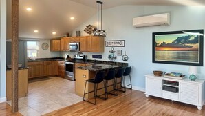 A modern kitchen with wooden cabinets, a central island with bar stools, a large window, and a wall-mounted picture of a sunset. - Top Mast Resort-Anchor House, Private Beach Access. (North Truro)