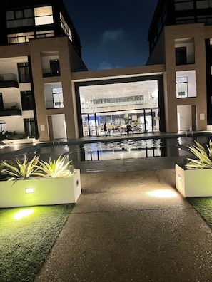 A modern building with a large glass entrance, outdoor seating area, and a reflective pool. - Embassy Gardens Royal Luxury Apartments (Accra)