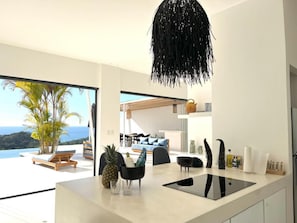 Une cuisine moderne dotée d’un grand îlot central, d’un luminaire suspendu et offrant une vue sur un balcon aménagé avec du mobilier d’extérieur.