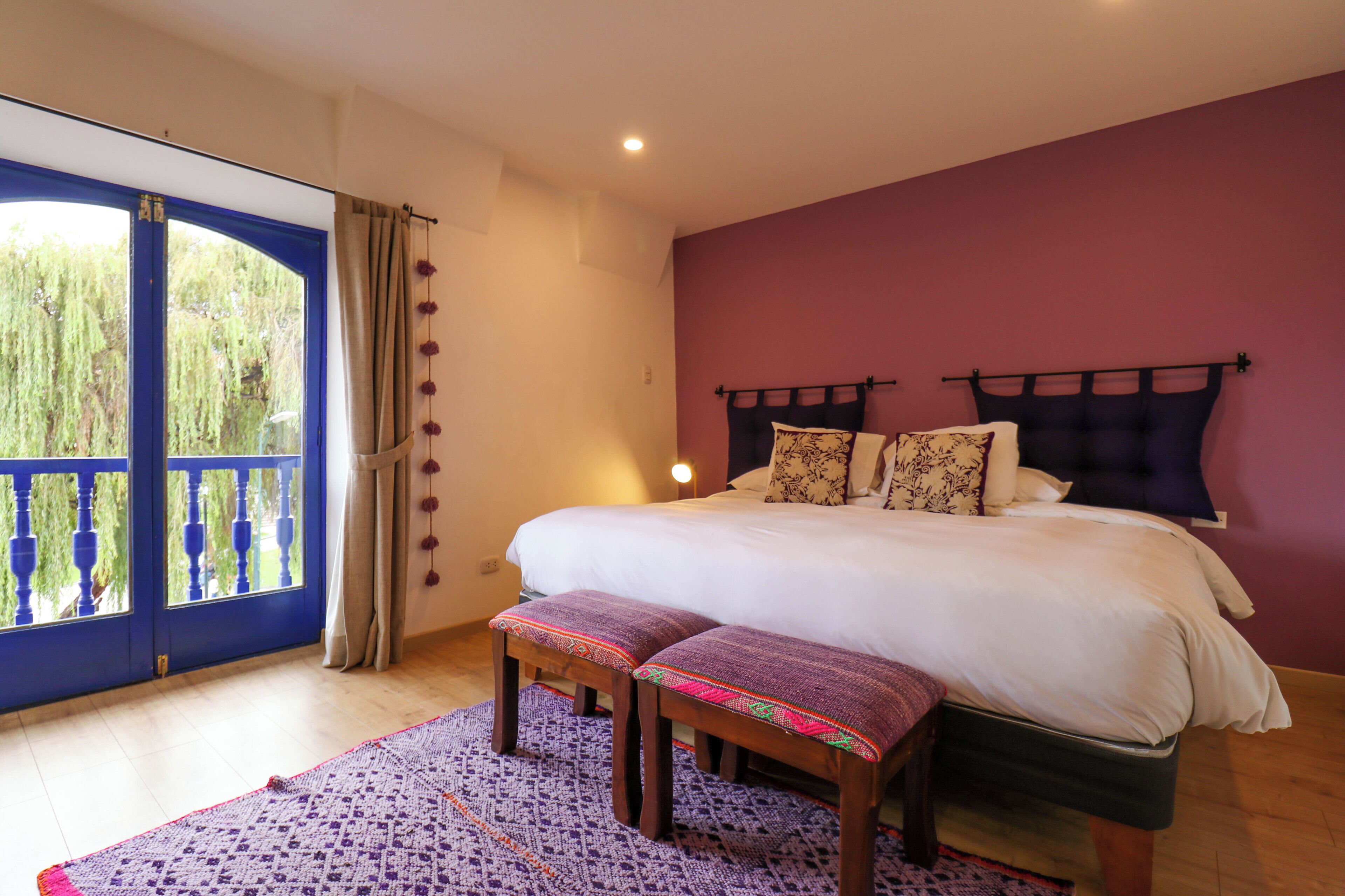 A bedroom with a large bed, a bench, a patterned rug, and a view of greenery through a blue-framed window.