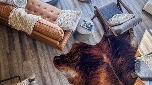 Une pièce chaleureuse avec une grande fenêtre, un canapé en cuir, un pouf rond, une chaise en bois et une grande moquette en fourrure.