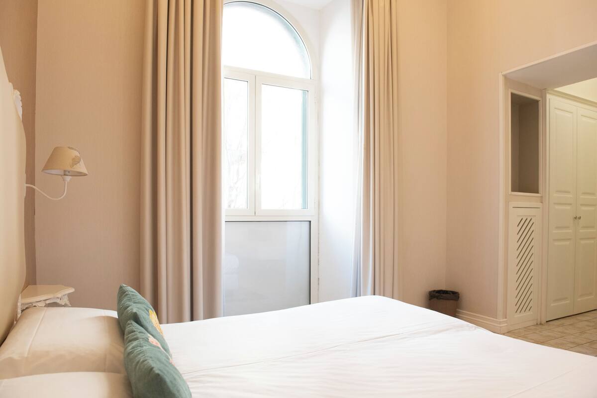 Una habitación de hotel con cama, mesita de noche, lámpara, ventana con cortinas y armario.