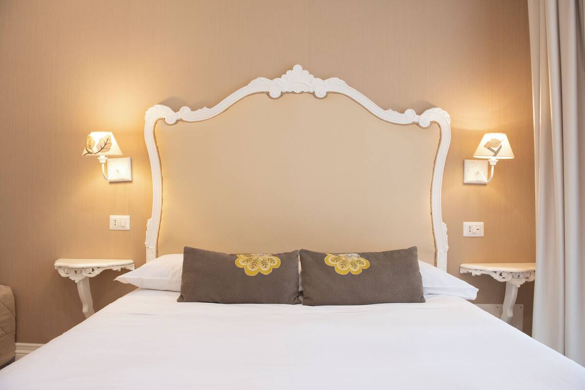 Una cama bien tendida con cabecera blanca, dos mesitas de noche con lámparas y una pared color crema.