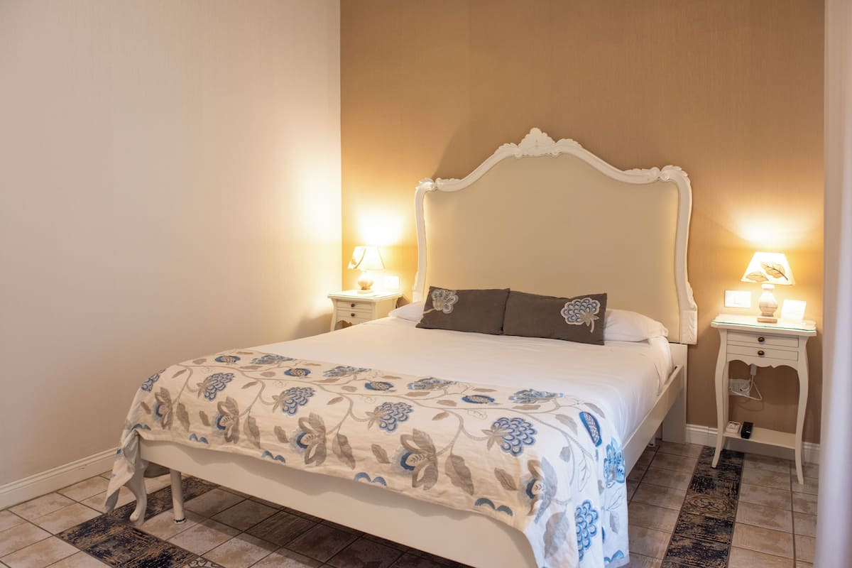 Una cama bien hecha con colcha estampada, dos mesitas de noche con lámparas y una alfombra decorada sobre suelo de baldosas.
