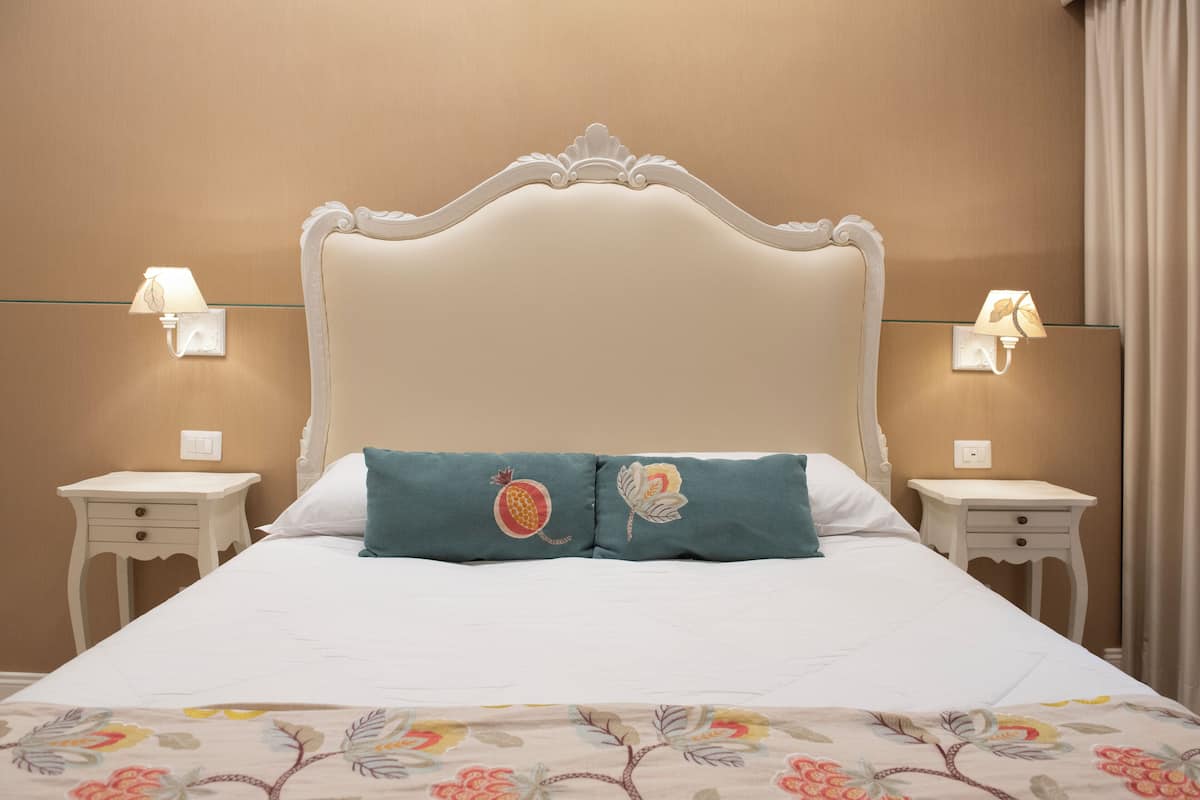 Una cama bien hecha con una colcha estampada con motivos florales, dos almohadas decorativas y dos mesitas de noche con lámparas.