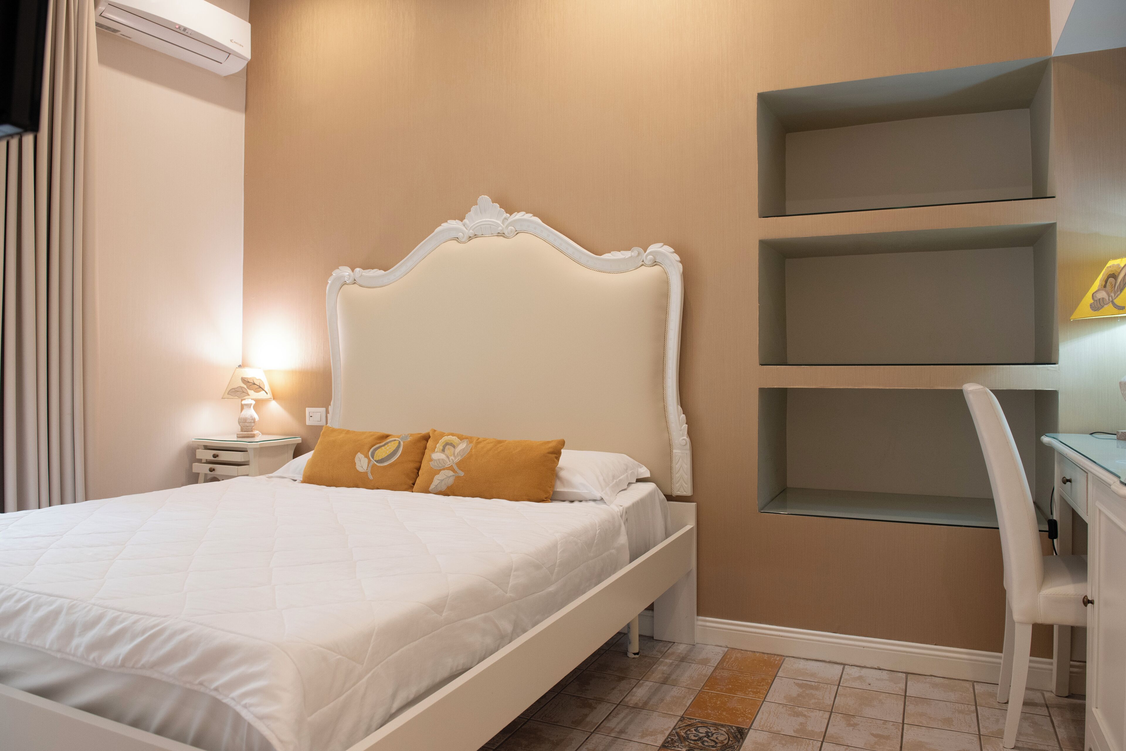 A neatly made bed with a white headboard, a bedside table with a lamp, and a chair with a desk against the wall.