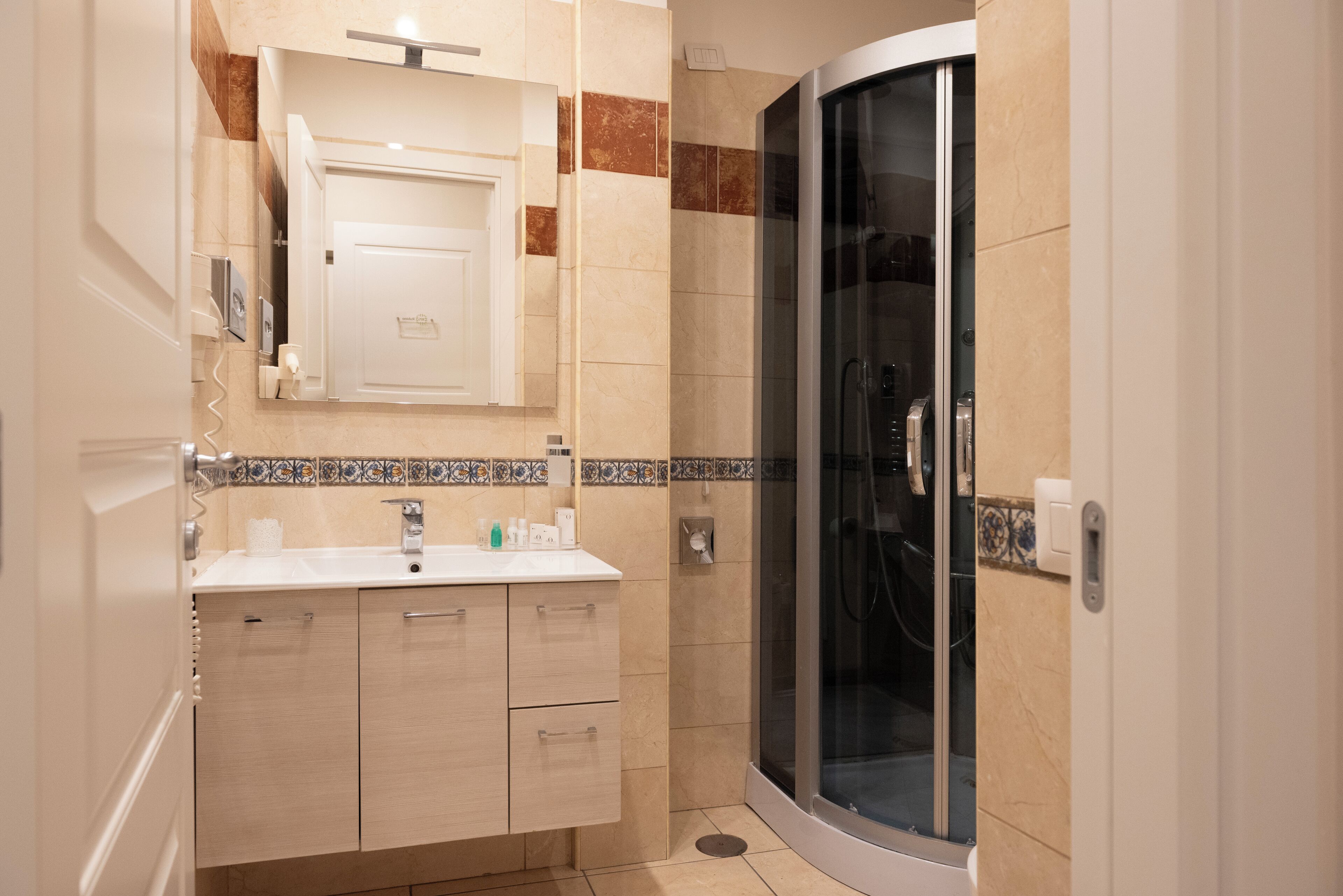A modern bathroom with a curved glass shower enclosure, a white sink with a mirror above, and beige tiled walls.