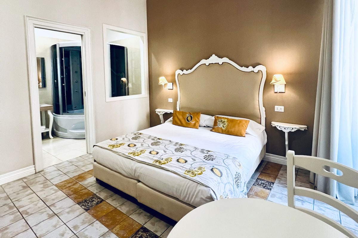 Un dormitorio con una cama grande, mesitas de noche con lámparas, una silla y vistas al baño.