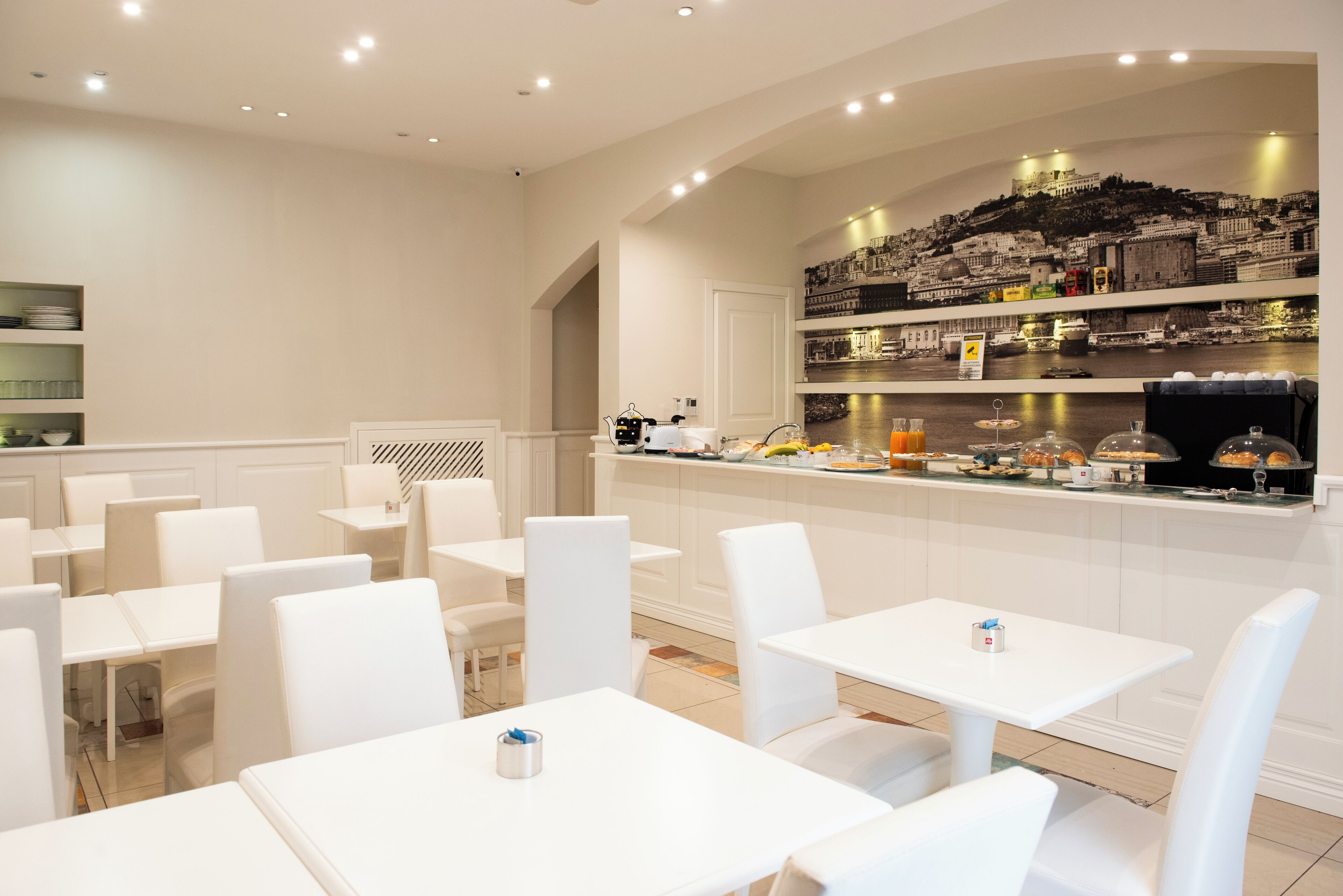 A restaurant with white tables and chairs, a counter with food displays, and a cityscape mural on the wall.