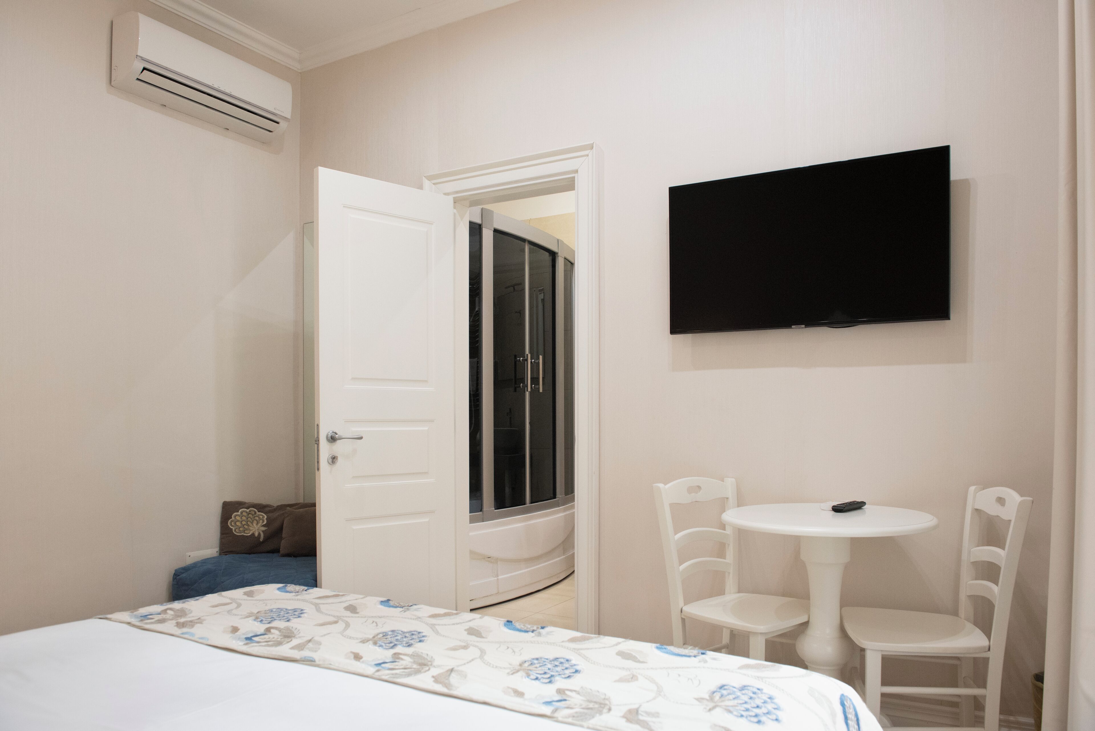 Un dormitorio con cama, televisor montado en la pared, una mesita con sillas y una puerta abierta que da a un baño.