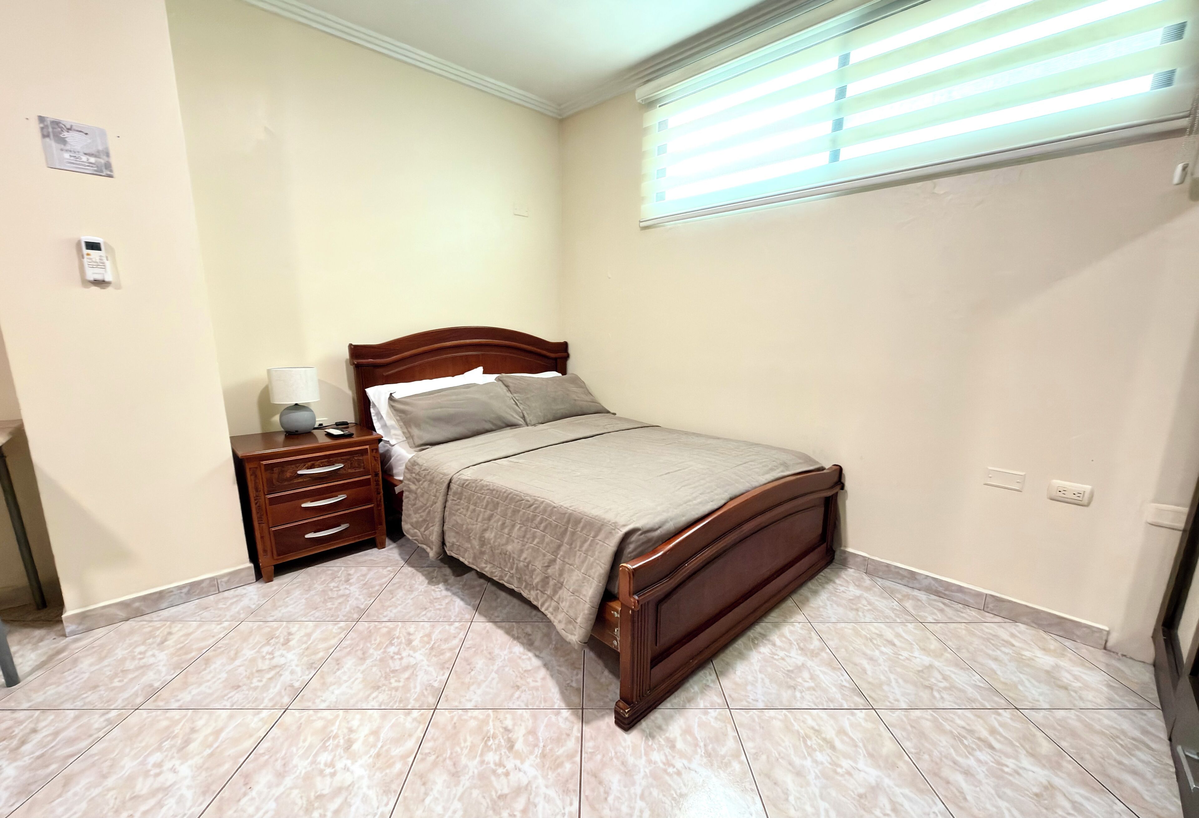 A bedroom with a bed, bedside table, lamp, and a window with blinds.
