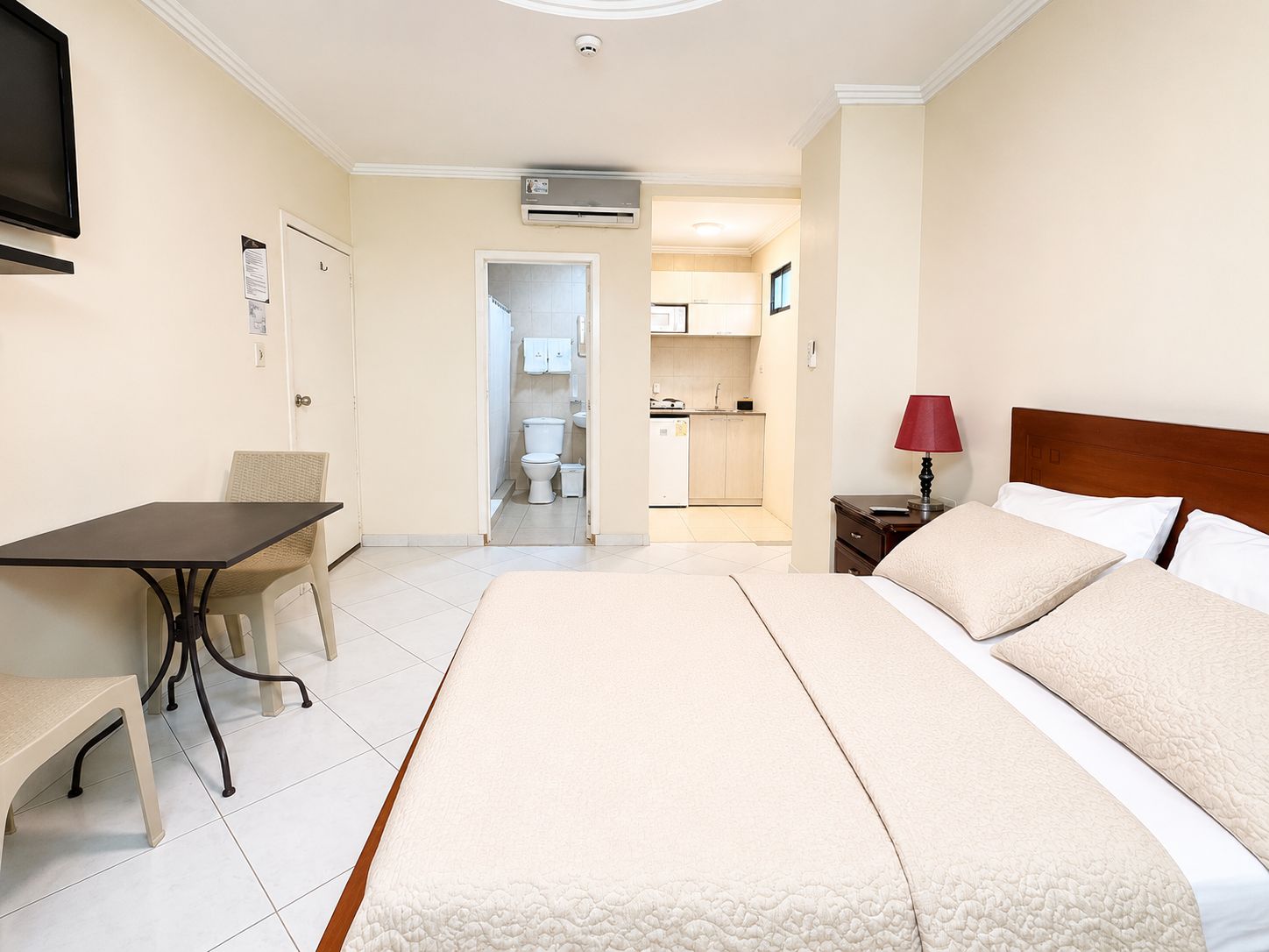 Une chambre d’hôtel comprenant un lit, un bureau, une chaise, une table de chevet, une lampe, et donnant sur la salle de bain et la cuisine.