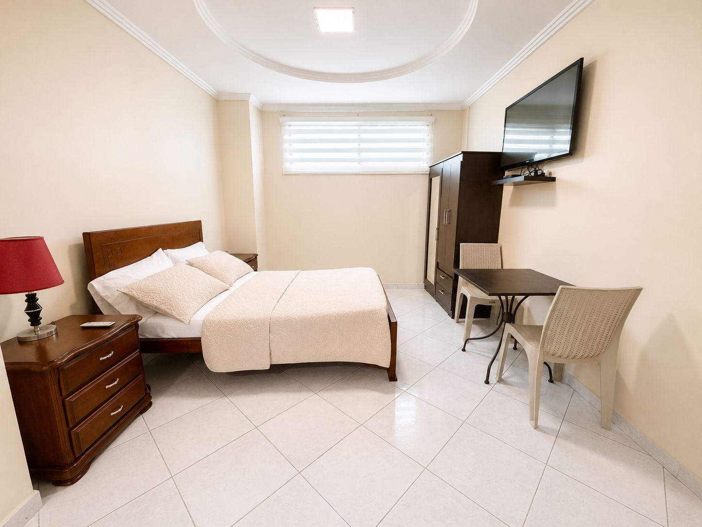A bedroom with a bed, bedside table, lamp, dresser, desk, chair, TV, and window with blinds.