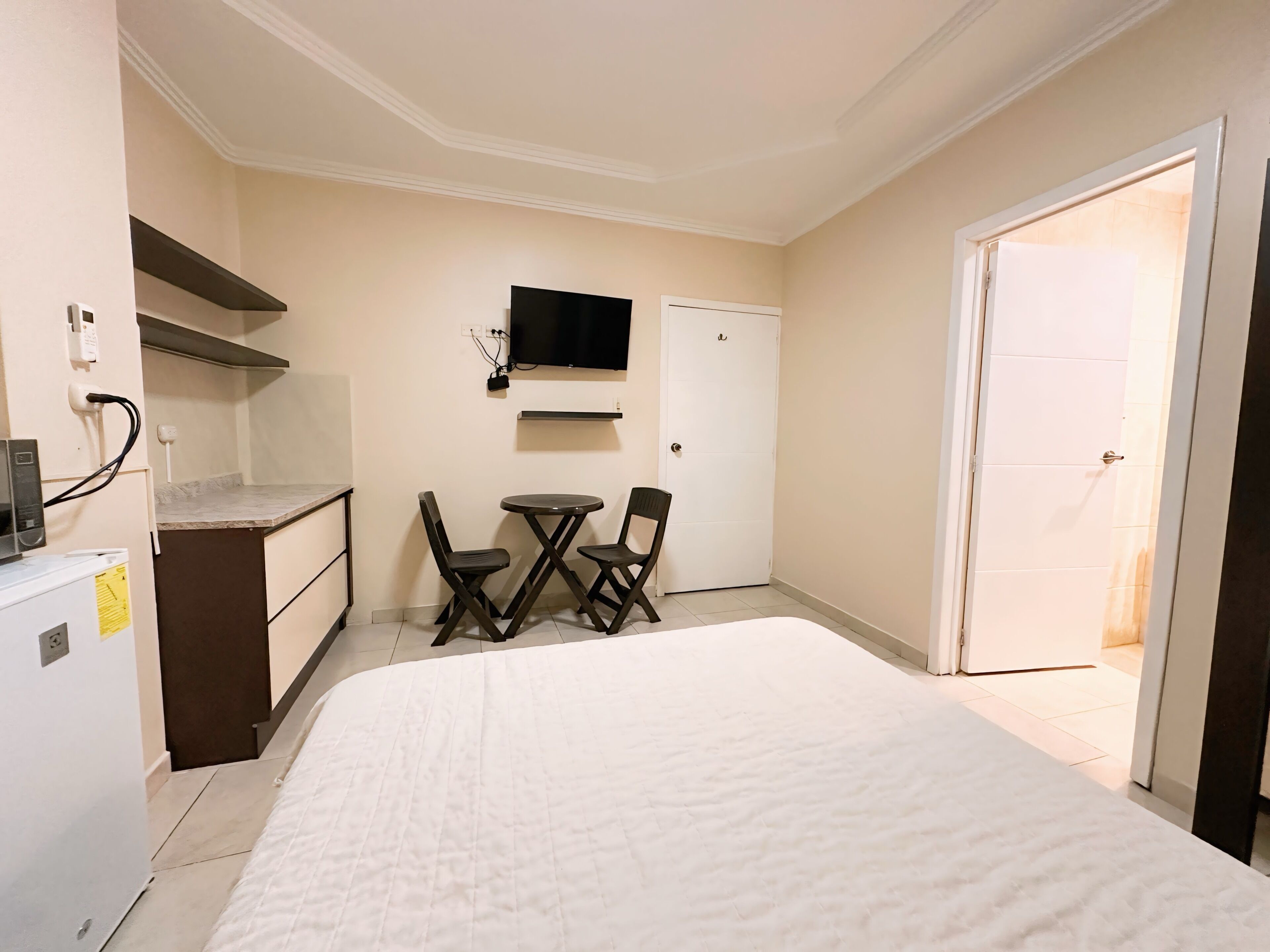 A hotel room with a bed, a small dining table with chairs, a TV mounted on the wall, and a door leading to another room.