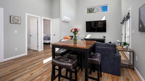 A modern living room with a wooden dining table, leather bar stools, a grey sofa, a flat-screen TV, and a small potted plant. - Hot Tub / Near Downtown / A/C / Couples Retreat (Colorado Springs)