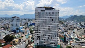 Un edificio de gran altura con varios pisos, rodeado de una zona urbana densamente poblada con diversos edificios y un paisaje montañoso al fondo.