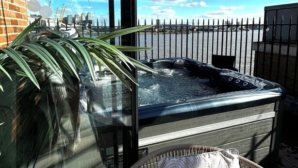 A hot tub with a glass enclosure, a wicker chair with cushions, and a view of a city skyline through a fence. - 3 Bdm Flat w/ HotTub and RoofTop (Greater London)