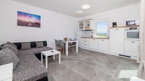 Una sala moderna con un sofá gris, una mesita blanca y un comedor con una mesa y sillas blancas. Se ve una cocina con armarios y electrodomésticos blancos al fondo.