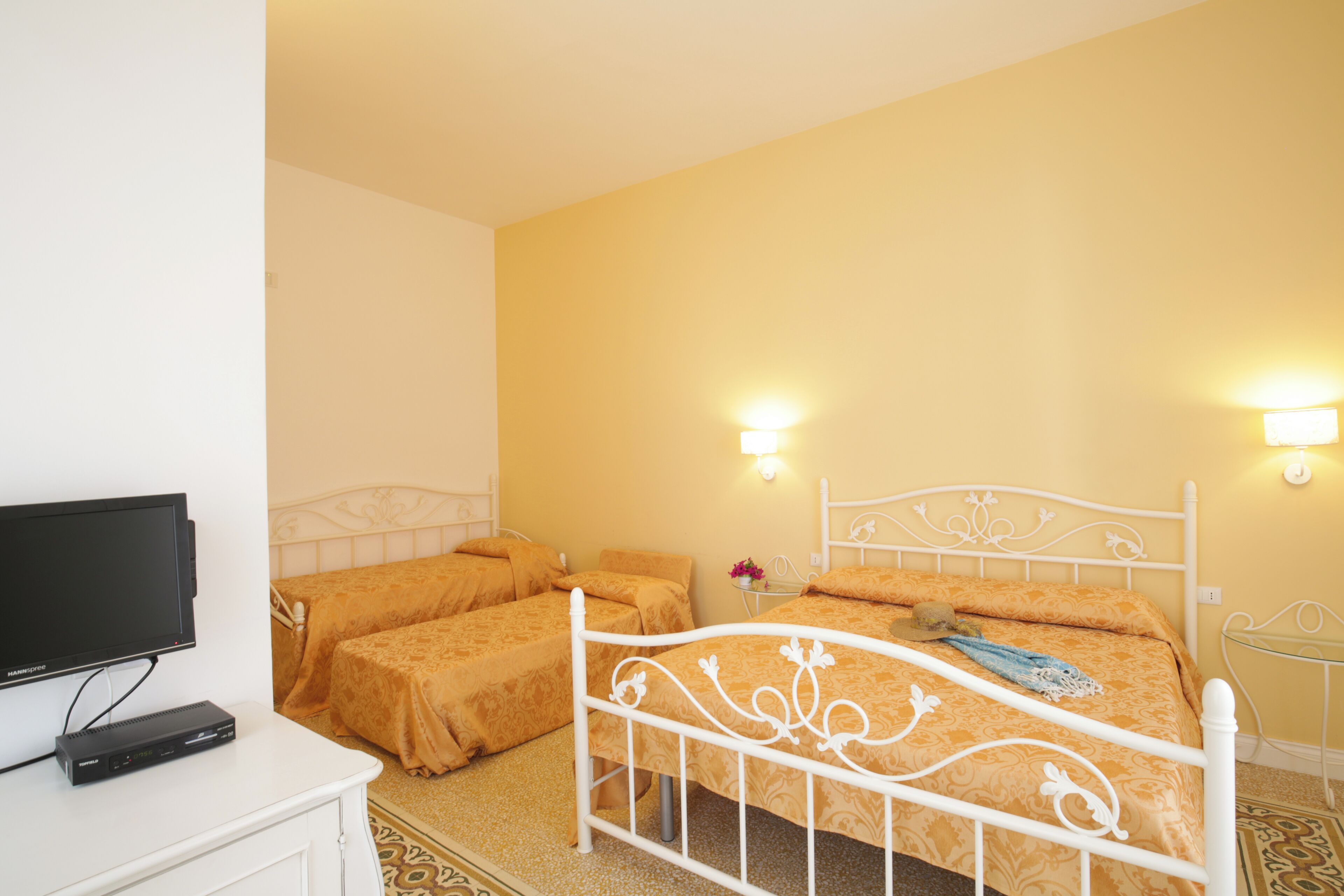 Une chambre d’hôtel avec un cadre de lit en métal blanc, deux lits avec la même literie orange, un téléviseur à écran plat posé sur une commode blanche et deux lampes fixées au mur.