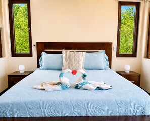 A bed with a blue quilt, a wooden headboard, two bedside tables with lamps, and windows with greenery outside. - Living Waters Casita  Private Pool, Camp Bay (Camp Bay)