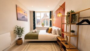 A bedroom with a bed, a window with curtains, a plant, a wooden shelf with clothes, and a radiator. - 3BR Westbourne Delight - Near Beach & Bournemouth! (Bournemouth)