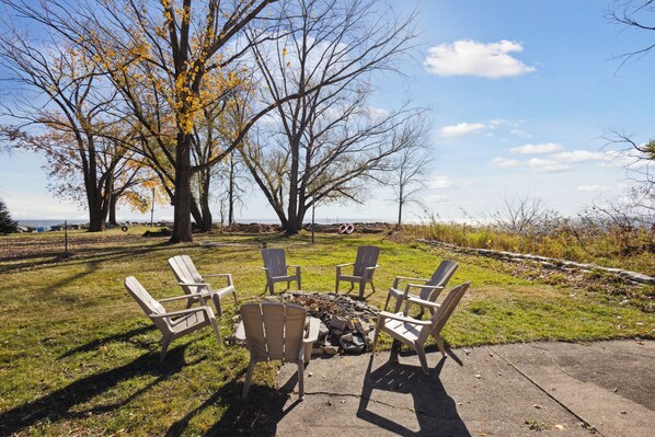 A fire pit with four Adirondack chairs arranged around it on a concrete patio, surrounded by grass and trees with autumn leaves.