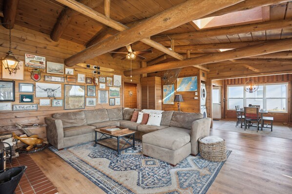 A cozy log cabin interior with a sectional sofa, wooden beams, framed pictures, and a dining area.