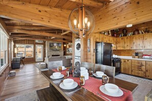A wooden interior with a dining area set for four, a kitchen with wooden cabinets, and a large window overlooking a garden.