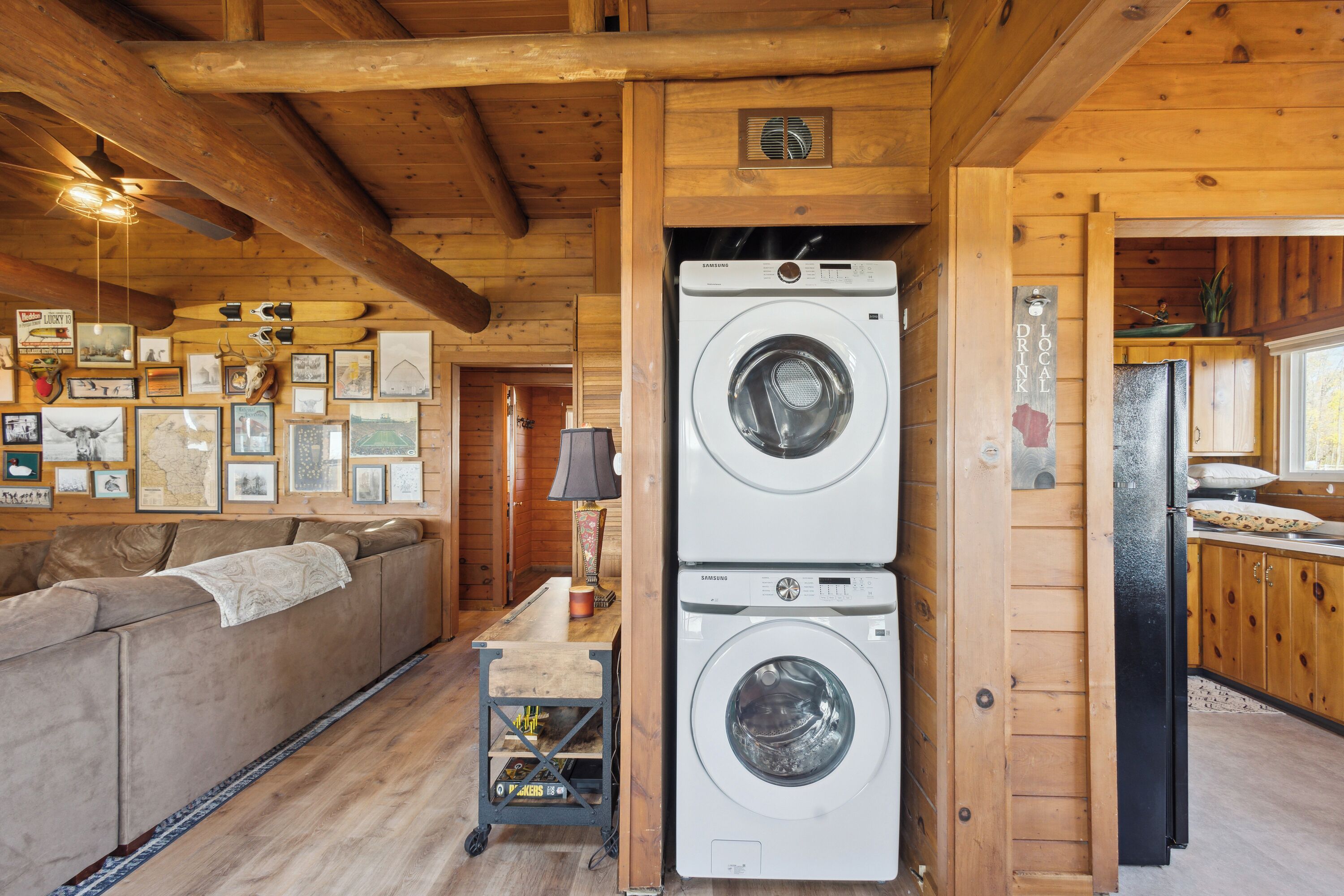 Interior acogedor de una cabaña con techo de madera, un sofá, una lavadora y un carrito con una lámpara.