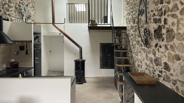 A spacious kitchen with a stone wall, a staircase, and a black countertop. - Maison de Caractère (Peyroules)