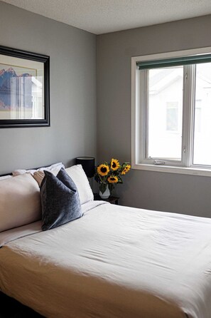 Un dormitorio con cama, almohadas, una mesita de noche, un florero con girasoles y una ventana.