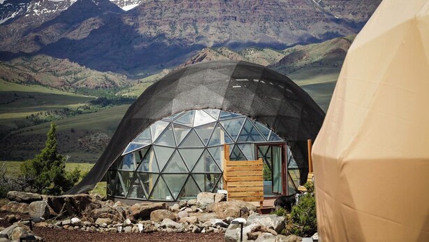 A geodesic dome structure with a wooden bench and a glass door, set against a mountainous backdrop. - Cody Peak Dome, Stunning Views! Near Cody, WY and Yellowstone National Park (Cody)