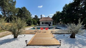 Une terrasse en bois menant à une maison dotée d’une piscine, le tout entouré d’arbres et de gravier.