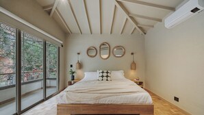 A bedroom with a large bed, wooden flooring, and a view of greenery through a sliding door.
