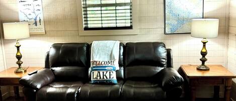 A living room with a leather couch, two lamps, a rug, a map on the wall, and a window with blinds.
