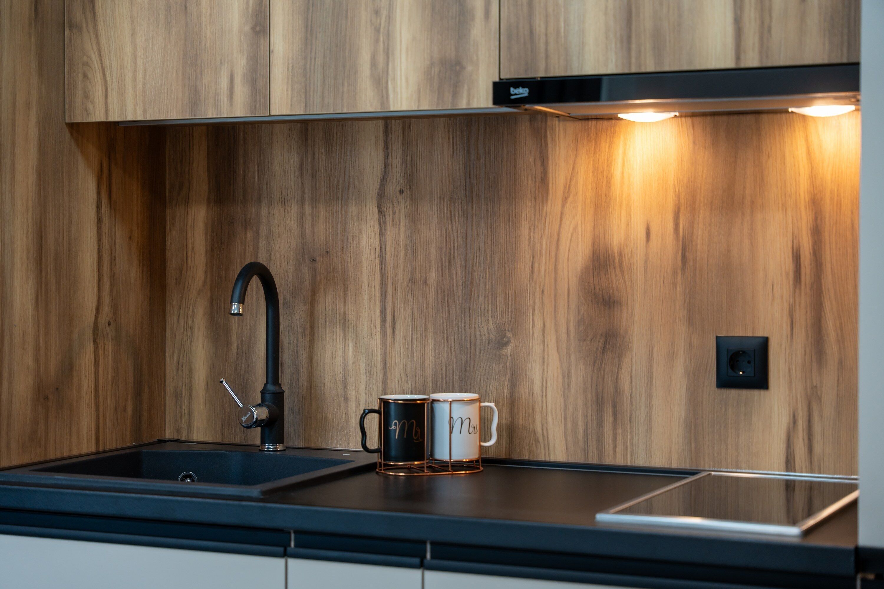 A modern kitchen with a wooden backsplash, a black faucet, and two mugs on the countertop.
