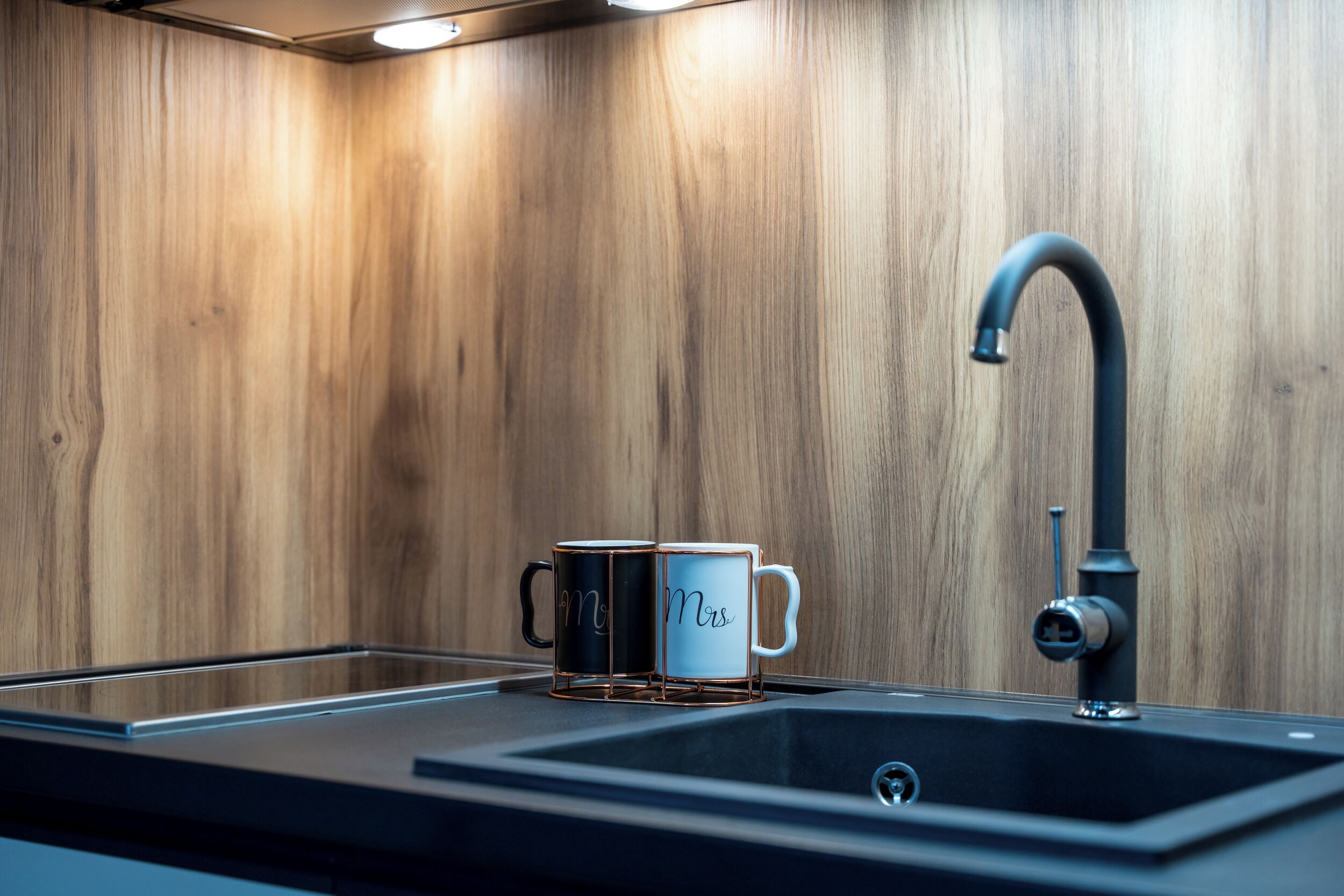 A modern kitchen with a wooden backsplash, a black sink, and two mugs with "Mr" and "Mrs" on them.