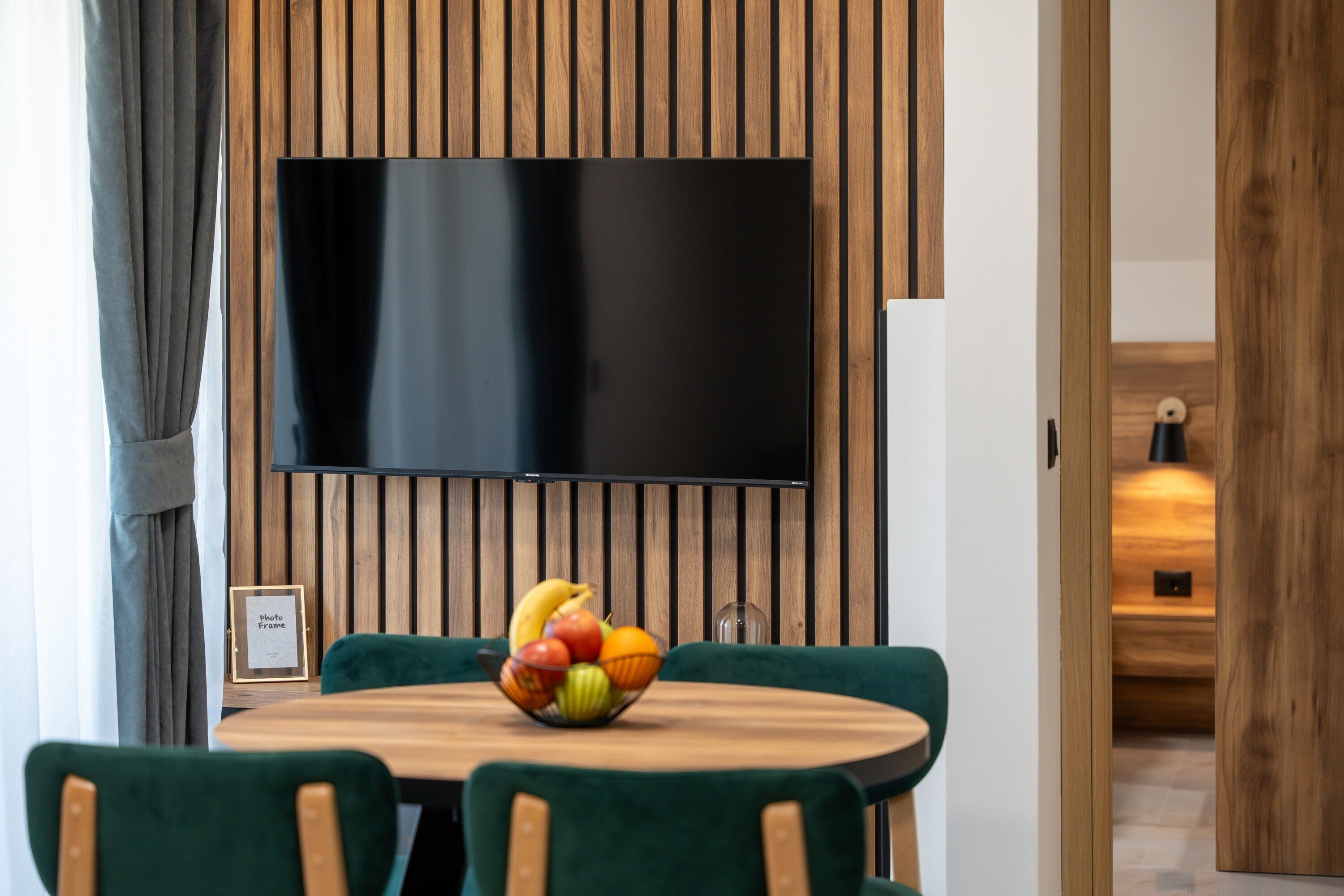 A dining area with a round wooden table, green upholstered chairs, and a fruit bowl. A flat-screen TV is mounted on the wall above the table.