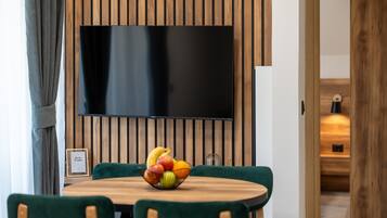 A dining area with a round wooden table, green upholstered chairs, and a fruit bowl. A flat-screen TV is mounted on the wall above the table.