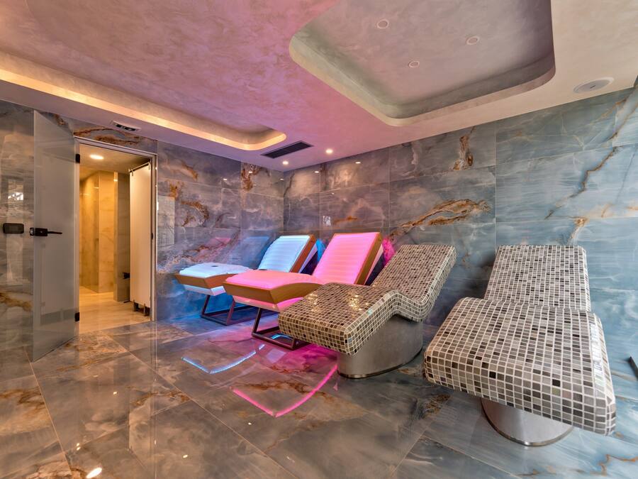 Un moderno spa con paredes y suelo de mármol, puerta de cristal y dos sillones de masaje con iluminación rosada.