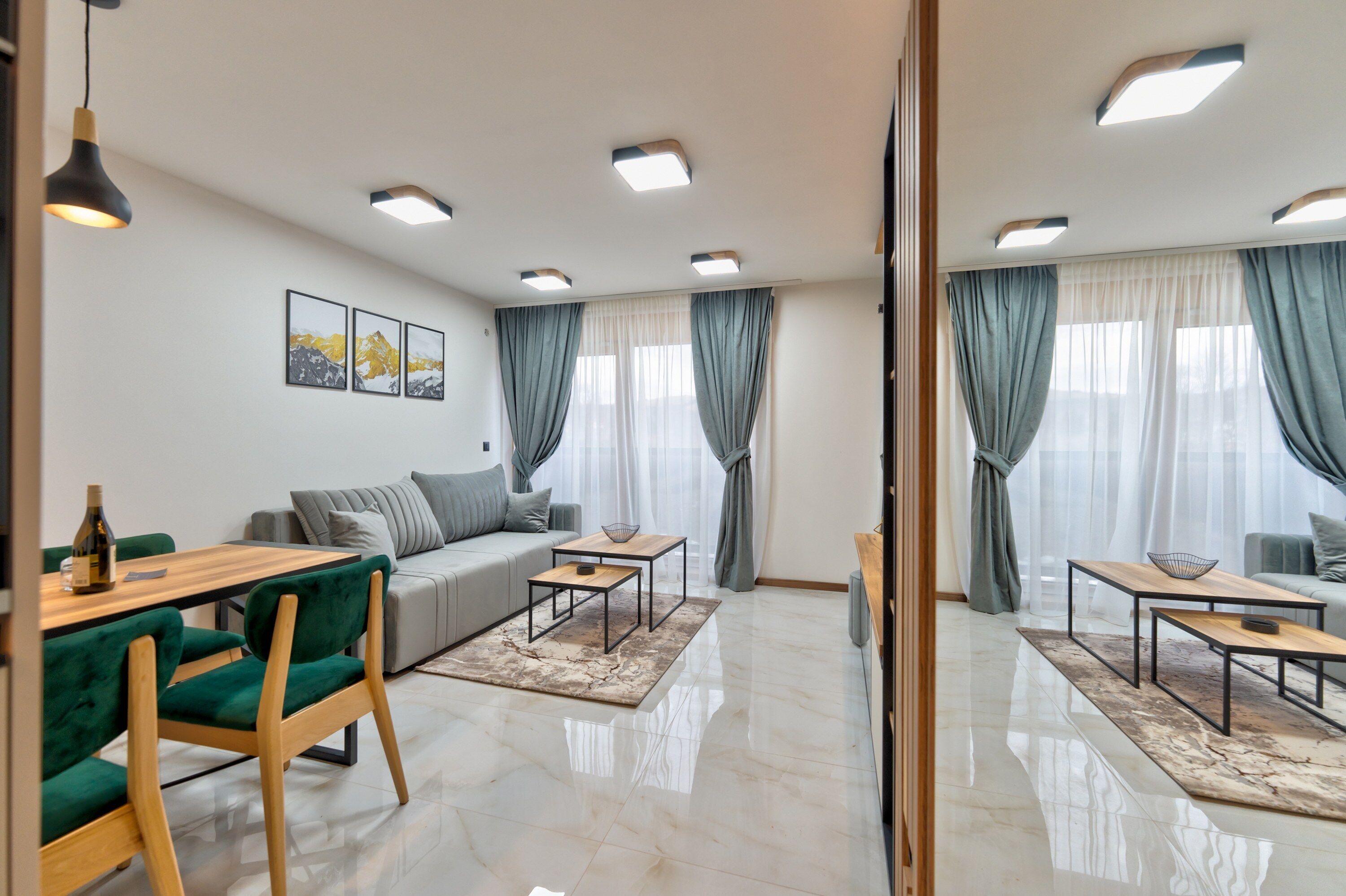 A modern living room with a sofa, a dining table, and a coffee table. There are ceiling lights and a large window with curtains.