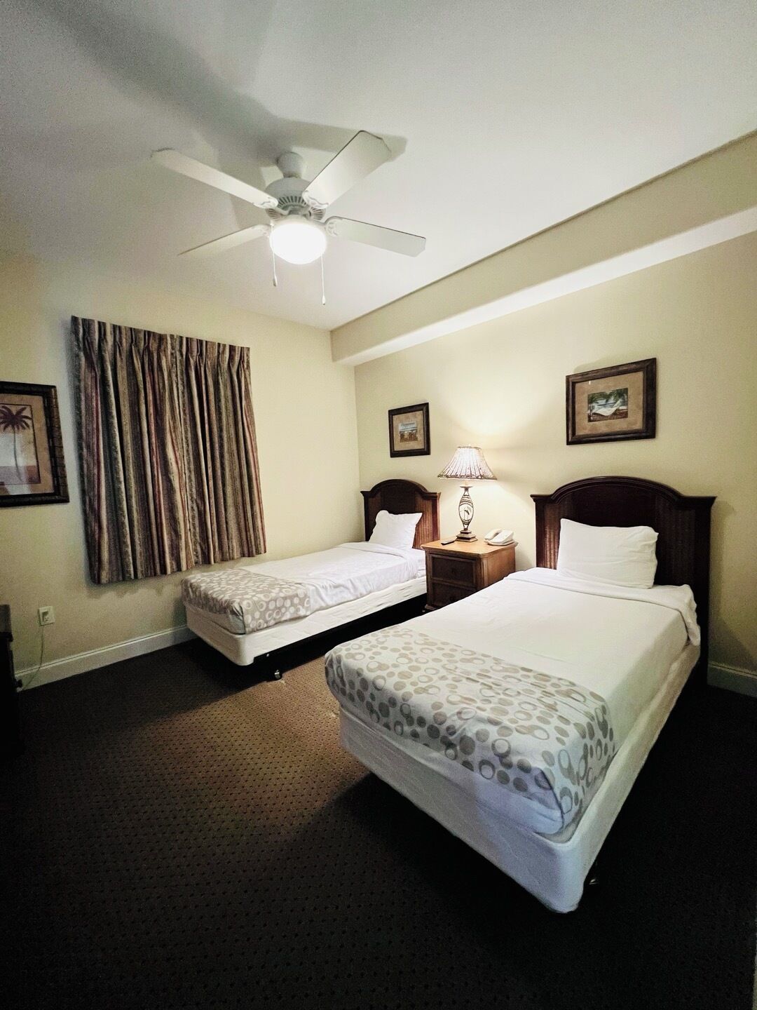 Een hotelkamer met twee bedden, een plafondventilator, een lamp en ingekaderde foto’s aan de muur.