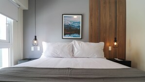 A neatly made bed with white linens, two bedside tables with lamps, a framed picture on the wall, and a window on the left.