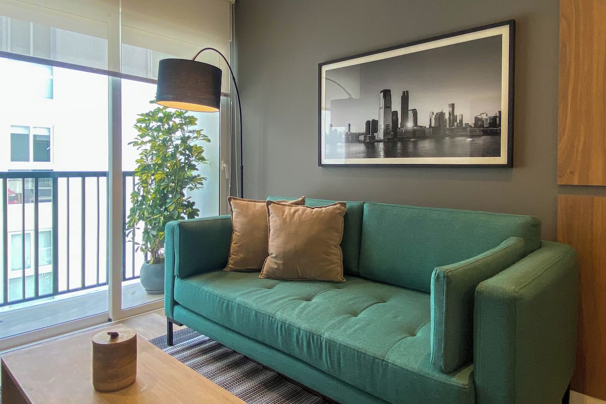 Een moderne woonkamer met een turquoise bank, een afbeelding van een stadsgezicht in een lijst, een vloerlamp en een kamerplant.