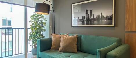 Una sala de estar moderna con un sofá color turquesa, un cuadro con vista a la ciudad, una lámpara de pie y una planta en maceta.