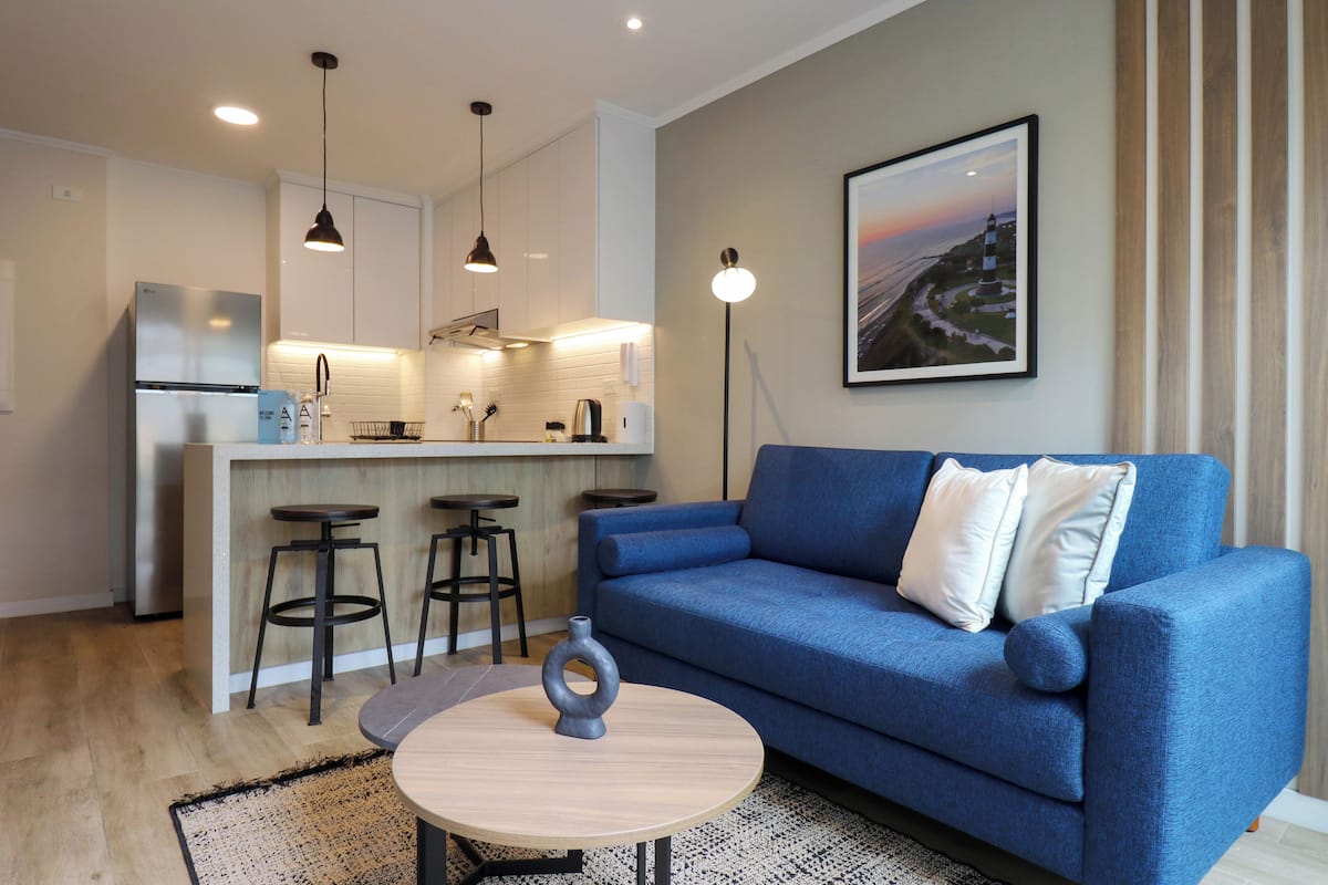 Een moderne woonkamer met een blauwe bank, een ronde salontafel en een keukenhoek met een barstoel.
